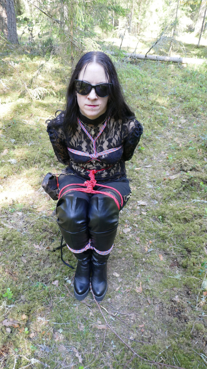 dark-haired female is tied to a tree in sun glasses and leather clothing
